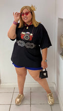 Load image into Gallery viewer, Woman in a black t-shirt with a camera design, blue shorts, and gold shoes standing in a boutique.

